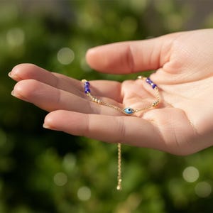 May include: A gold-tone chain bracelet featuring a central blue and white evil eye charm, accented with small gold-tone beads and deep blue spherical beads along the chain, finished with a delicate adjustable extender chain.