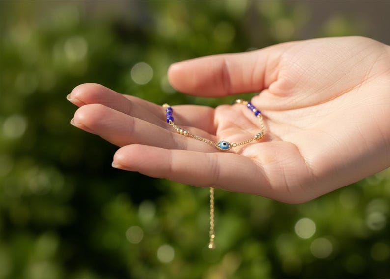 May include: A gold-tone chain bracelet featuring a central blue and white evil eye charm, accented with small gold-tone beads and deep blue spherical beads along the chain, finished with a delicate adjustable extender chain.