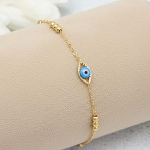 May include: A gold-toned chain bracelet featuring a central eye-shaped charm with a blue, white, and black pupil design. The bracelet includes small gold-toned spherical beads spaced along the delicate metal chain links for a refined appearance.