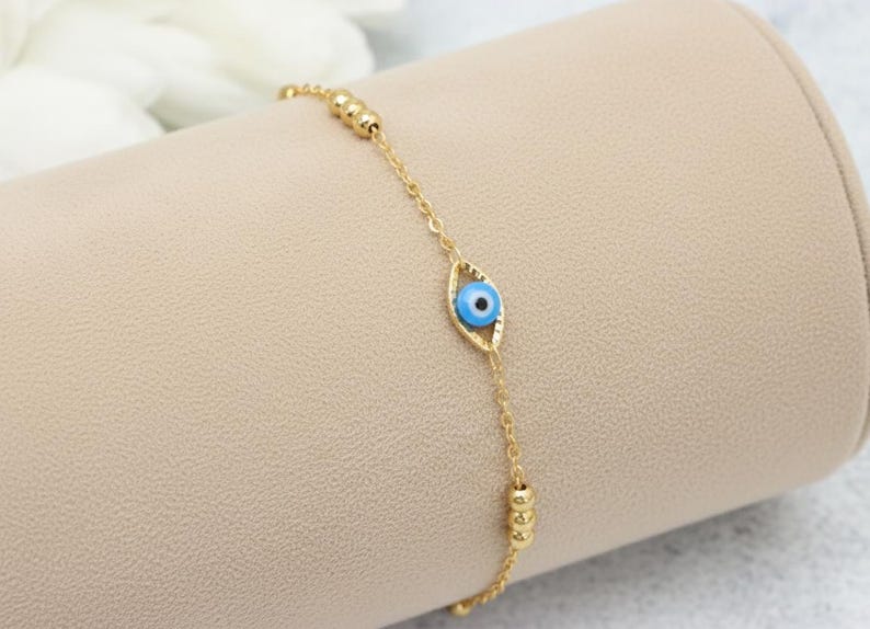 May include: A gold-toned chain bracelet featuring a central eye-shaped charm with a blue, white, and black pupil design. The bracelet includes small gold-toned spherical beads spaced along the delicate metal chain links for a refined appearance.