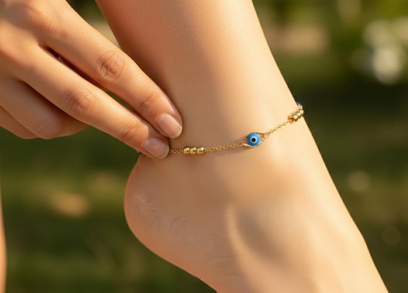 May include: A gold-toned chain anklet featuring a central blue and white evil eye charm. The delicate metal chain is accented with small, round gold-colored beads spaced along the length, creating a simple and refined piece of jewelry.