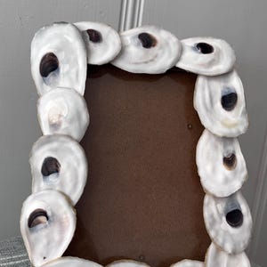 May include: A rectangular photo frame decorated with white oyster shells. The shells are arranged around the brown frame, creating a coastal or nautical aesthetic. The shells have dark brown and black interiors.
