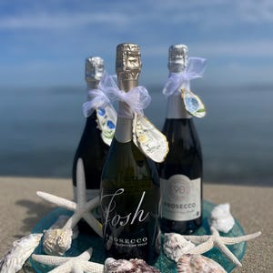 May include: Three Prosecco bottles with silver foil tops and white bows, on a blue glass plate with starfish and seashells. The bottles are labelled "Josh Prosecco Product of Italy". The background shows the sea and sky.