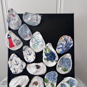 May include: A display of painted oyster shells on a black stand. The shells are decorated with maps, anchors, and floral designs in blue, white, and red. Some shells have the text "Cape Cod". The designs are in a variety of styles.