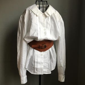 Pu&ograve; includere: Una camicia bianca a maniche lunghe con bottoni, colletto grande e cintura in pelle marrone.