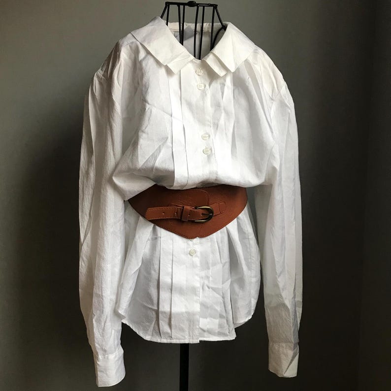 Pu&ograve; includere: Una camicia bianca a maniche lunghe con bottoni, colletto grande e cintura in pelle marrone.
