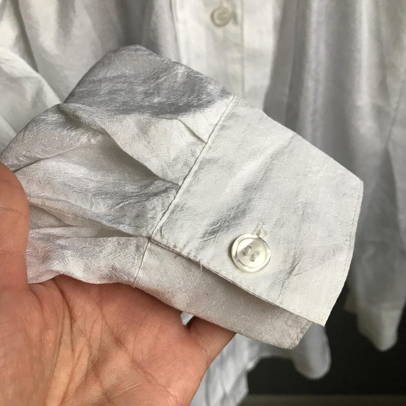 Pu&ograve; includere: Camicia bianca in seta con bottoni, con un tessuto strutturato e stropicciato. La camicia ha un polsino con un bottone e una chiusura con un bottone al collo.