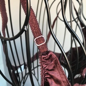 May include: A burgundy bra with adjustable straps and a silver metal clasp. The bra is on a black wire hanger.