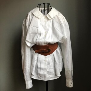 Pu&ograve; includere: Una camicia bianca oversize con bottoni e una cintura in pelle marrone. La camicia ha un colletto classico e maniche lunghe.