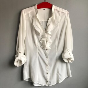 May include: White long-sleeved blouse with a ruffled collar and button closure.