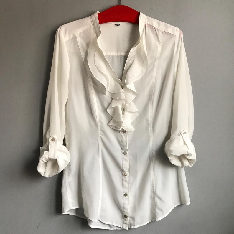 May include: White long-sleeved blouse with a ruffled collar and button closure.