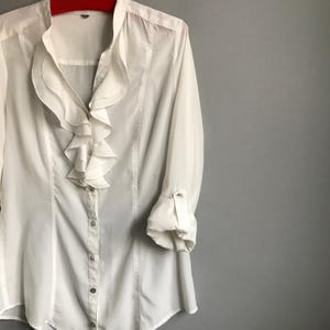 May include: A white long-sleeved blouse with a ruffled collar and button-up front.