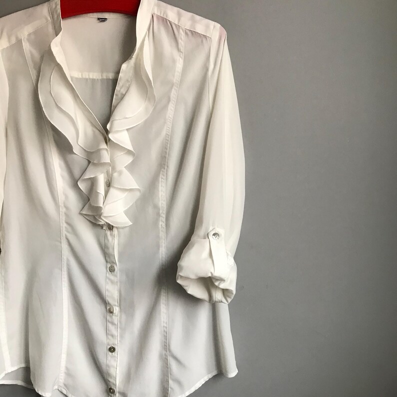 May include: A white long-sleeved blouse with a ruffled collar and button-up front.