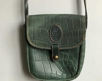 May include: A green leather crossbody bag with a flap closure and a metal buckle. The bag has a crocodile-embossed pattern and a logo patch that reads "Scotch & Soda".