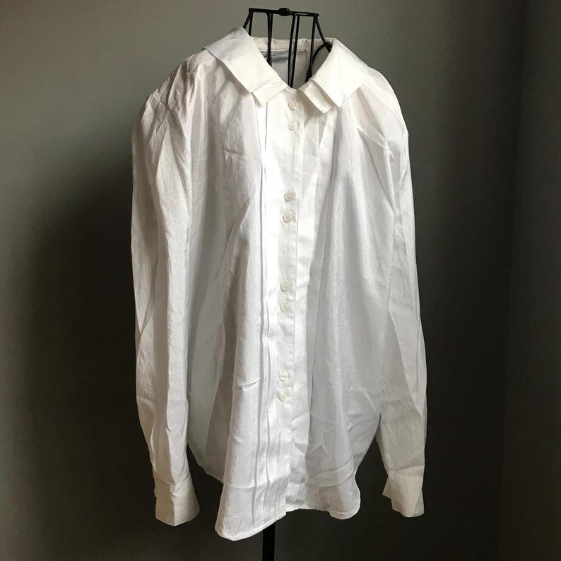 Pu&ograve; includere: Una camicia bianca a maniche lunghe con bottoni e colletto. La camicia ha una vestibilit&agrave; rilassata e una silhouette leggermente oversize.