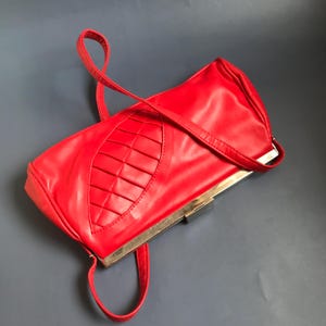 Op de afbeelding: Een rode leren clutch met een zilveren sluiting en een lange band. De clutch heeft een uniek ontwerp met een geweven patroon aan de voorkant.