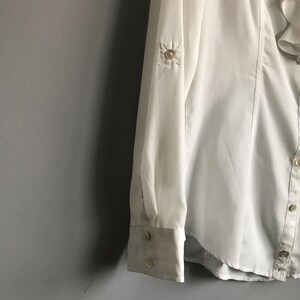 May include: White long-sleeve button-up shirt with a ruffled collar and a small button detail on the sleeve.