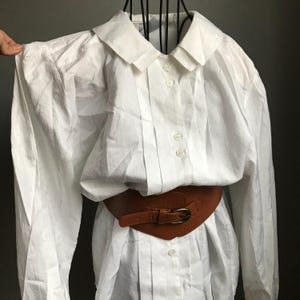 Pu&ograve; includere: Una camicia bianca a maniche lunghe con colletto alla Peter Pan e una cintura in pelle marrone. La camicia ha una chiusura con bottoni sul davanti ed &egrave; realizzata in un tessuto leggero.