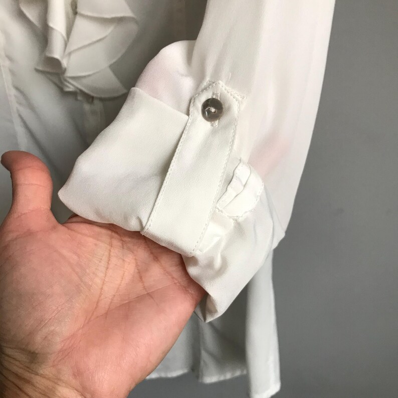 May include: White long-sleeve blouse with a ruffled neckline and a button detail on the cuff.