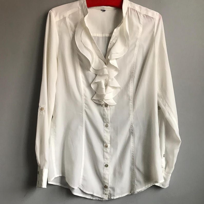 May include: White long-sleeved button-up blouse with a large ruffled collar.