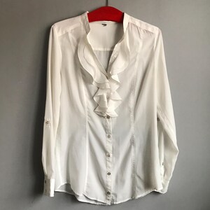 May include: A white long-sleeved button-up shirt with a large ruffled collar.