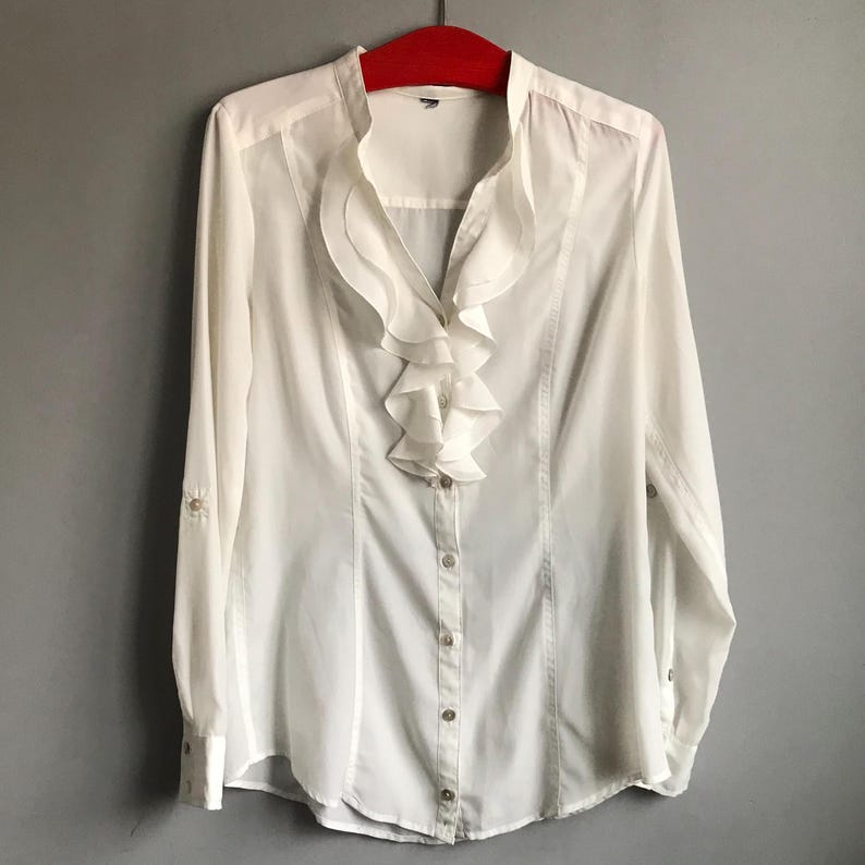 May include: A white long-sleeved button-up shirt with a large ruffled collar.