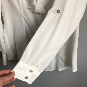 May include: White long-sleeve blouse with a ruffled collar and button closures. The blouse has a white button on the front and two gold buttons on the cuff.