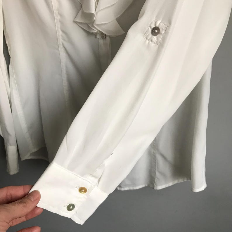 May include: White long-sleeve blouse with a ruffled collar and button closures. The blouse has a white button on the front and two gold buttons on the cuff.