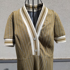 May include: A vintage-style dress in a mustard yellow hue, featuring a V-neckline with white and olive green stripes. The dress has short sleeves with matching striped cuffs and is adorned with small blue dots. Two buttons secure the front.