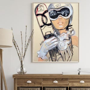 May include: A framed art print featuring a stylized illustration of a person wearing a ski helmet and goggles with a Chanel logo. The person holds a snowboard with a prominent Chanel logo. The color palette includes cream, black, and white tones.