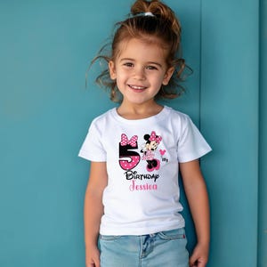 Op de afbeelding: Een wit T-shirt met een Minnie Mouse-ontwerp. Het ontwerp bevat het nummer 5 in roze stippen, de tekst "Birthday Jessica" en een Minnie Mouse-afbeelding. Het shirt wordt gedragen door een jong meisje.
