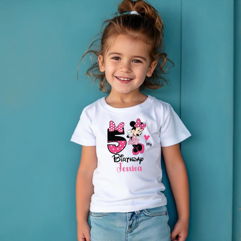 Op de afbeelding: Een wit T-shirt met een Minnie Mouse-ontwerp. Het ontwerp bevat het nummer 5 in roze stippen, de tekst "Birthday Jessica" en een Minnie Mouse-afbeelding. Het shirt wordt gedragen door een jong meisje.