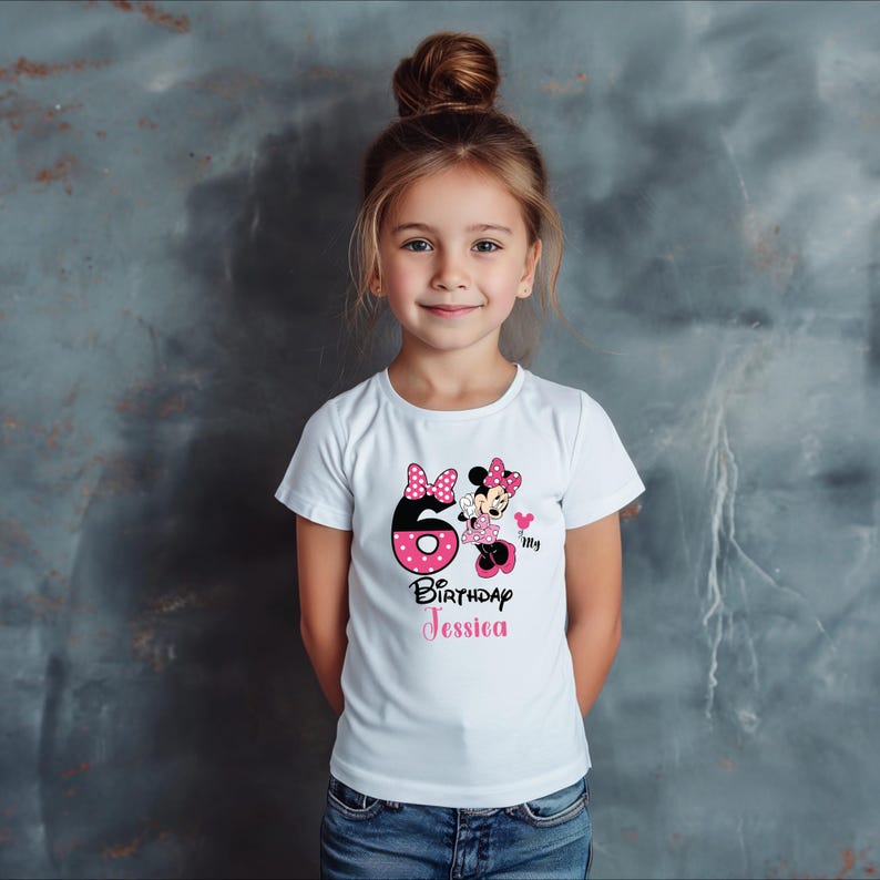 Op de afbeelding: Wit T-shirt met een Minnie Mouse-ontwerp, een roze strik met stippen en het nummer 6. De tekst "Birthday Jessica" is in het roze gedrukt. Het shirt is voor een kind.