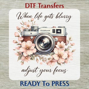 May include: DTF Transfers design featuring a vintage camera illustration with floral accents. The image includes the text "When life gets blurry adjust your focus" and "READY To PRESS". The camera is brown and silver, surrounded by pink flowers.