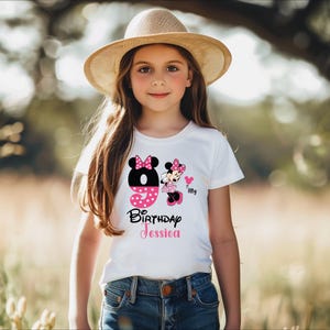 Op de afbeelding: Een wit T-shirt met een Minnie Mouse-verjaardagsontwerp. Het ontwerp bevat het nummer 9, een Minnie Mouse-afbeelding en de tekst "Birthday Jessica". Het shirt wordt gedragen door een jong meisje dat een strohoed draagt.