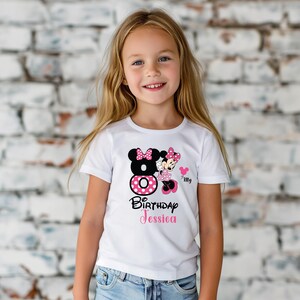 Op de afbeelding: Wit T-shirt met een Minnie Mouse-ontwerp voor een 8e verjaardag. Het ontwerp bevat een zwart nummer 8 met een roze stippelcirkel, Minnie Mouse en de naam "Jessica" in roze. Een roze hart is ook inbegrepen.