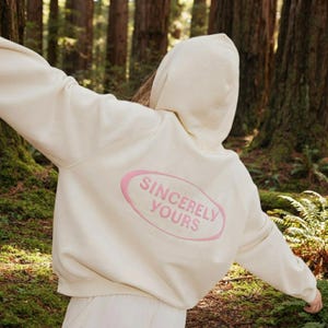 Puede incluir: Una sudadera con capucha de color crema que presenta un gráfico ovalado rosa en la parte posterior con el texto SINCERELY YOURS impreso en letras mayúsculas rosas. La prenda tiene mangas largas y capucha, mostrando un diseño sencillo y claro.