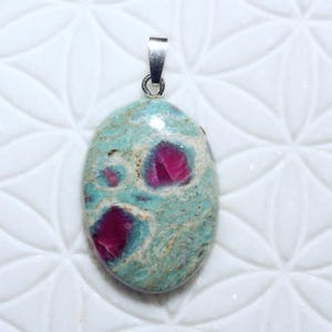 Könnte beinhalten: Ovaler Anhänger mit silberner Öse. Der Anhänger zeigt einen türkis-beigen Stein mit leuchtend magentafarbenen Einschlüssen. Der Stein hat eine polierte Oberfläche und ist vor einem weißen, strukturierten Hintergrund platziert. Der Text "B7316" befindet sich in der oberen linken Ecke.