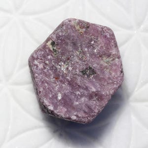 Könnte beinhalten: Ein roher, sechseckiger Rubin-Edelstein mit einer texturierten, purpurrosa Oberfläche und kleinen dunklen mineralischen Einschlüssen. Der Stein weist natürliche kristalline Facetten auf und ist mit dem alphanumerischen Code B9200 oben links versehen.