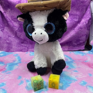 May include: A plush cow toy with black and white fur, large blue eyes, and a brown cowboy hat. Two small, square dice with embedded fruit slices and the number 3 and 6 are in front of the cow.