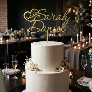Op de afbeelding: Een goudkleurige taarttopper met de namen Sarah en Daniel in cursief schrift, met een hartvorm die de datum 09.09.26 bevat. De topper staat bovenop een witte taart met twee lagen, versierd met kleine witte bloemen.