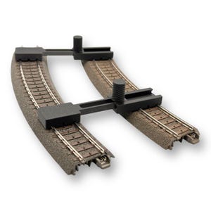 Könnte beinhalten: Ein gebogener Modelleisenbahn-Gleisabschnitt mit einem schwarzen Kunststoffverbinder. Die Strecke hat braunes Schotterbett und silberne Schienen. Der Verbinder hat zylindrische Pfosten und rechteckige Abschnitte. Weißer Hintergrund.