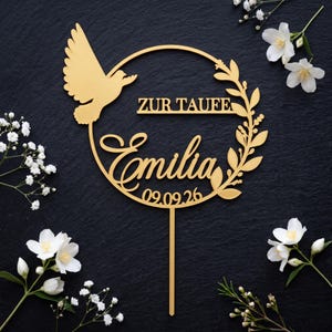 Può includere: Un topper per torta color oro caratterizzato da una cornice circolare con una colomba a sinistra, un ramo frondoso a destra e il testo "ZUR TAUFE", "Emilia" e "09.09.26" al centro, sostenuto da un singolo stelo verticale alla base.