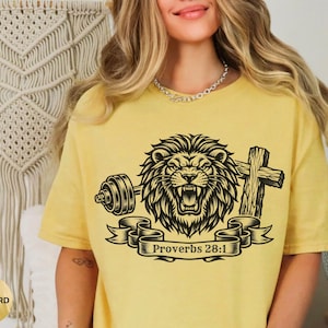 May include: Mustard yellow t-shirt featuring a black graphic of a roaring lion with a barbell, a cross, and a banner with the text "Proverbs 28:1". The shirt has short sleeves and a relaxed fit.