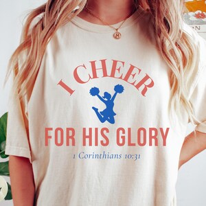 May include: Ivory-colored t-shirt with the text "I CHEER FOR HIS GLORY" in coral lettering. A blue cheerleader graphic is in the center. The Bible verse 1 Corinthians 10:31 is printed below the main text.