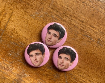 May include: Three pink button pins with a photo of a man's face on each. The man has dark hair and is wearing a white shirt.