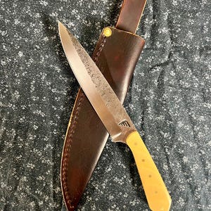 May include: A handcrafted chef's knife with a light-colored handle and a textured blade, resting on a dark brown leather sheath. The knife has a full tang construction and is designed for kitchen use.