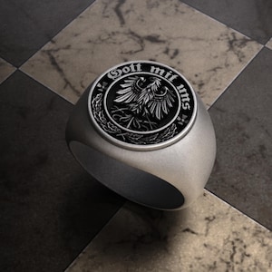 May include: A silver ring with a circular emblem featuring a detailed eagle design and the words "Gott mit uns". The ring has a brushed metal finish and a classic signet style.