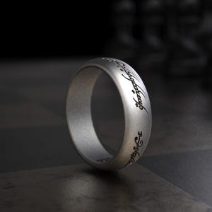 May include: A silver ring with engraved script. The ring has a matte finish and a simple band design. The script is in a stylized font and wraps around the ring. The ring is displayed on a dark surface.