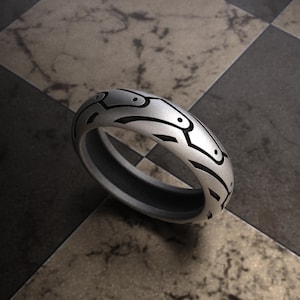 May include: A silver ring with a geometric design. The ring has a dark grey interior and a patterned exterior with black accents. The ring is set against a checkered background.